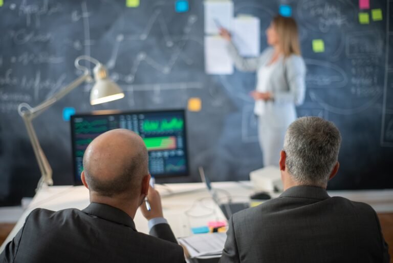 back-view-company-experts-working-office-two-serious-man-formalwear-sitting-looking-woman-presenting-business-plan-market-data-board-financial-analytics-business-growth-concept-min